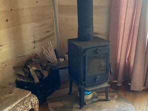 A wood-burning stove, a stack of firewood, and a window with curtains.