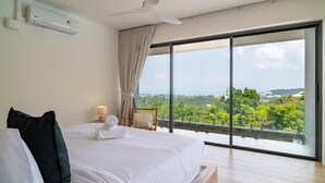 Une chambre dotée d’un grand lit, d’un ventilateur de plafond, d’une table de chevet avec une lampe, et offrant une vue sur la verdure et les bâtiments grâce à de grandes fenêtres.