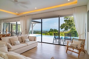 A spacious living room with a large sofa, a wicker chair, and a view of a swimming pool and greenery. - Brand New 3 bed Sea View Villa - KBR31 (Koh Samui)