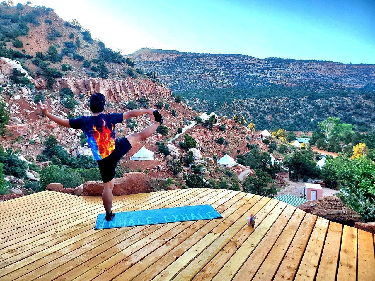 Une personne effectue une posture de yoga sur une terrasse en bois, avec en arrière-plan un paysage montagneux et des yourtes.