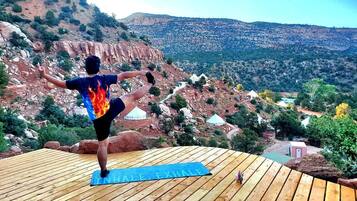 Une personne effectue une posture de yoga sur une terrasse en bois, avec en arrière-plan un paysage montagneux et des yourtes.