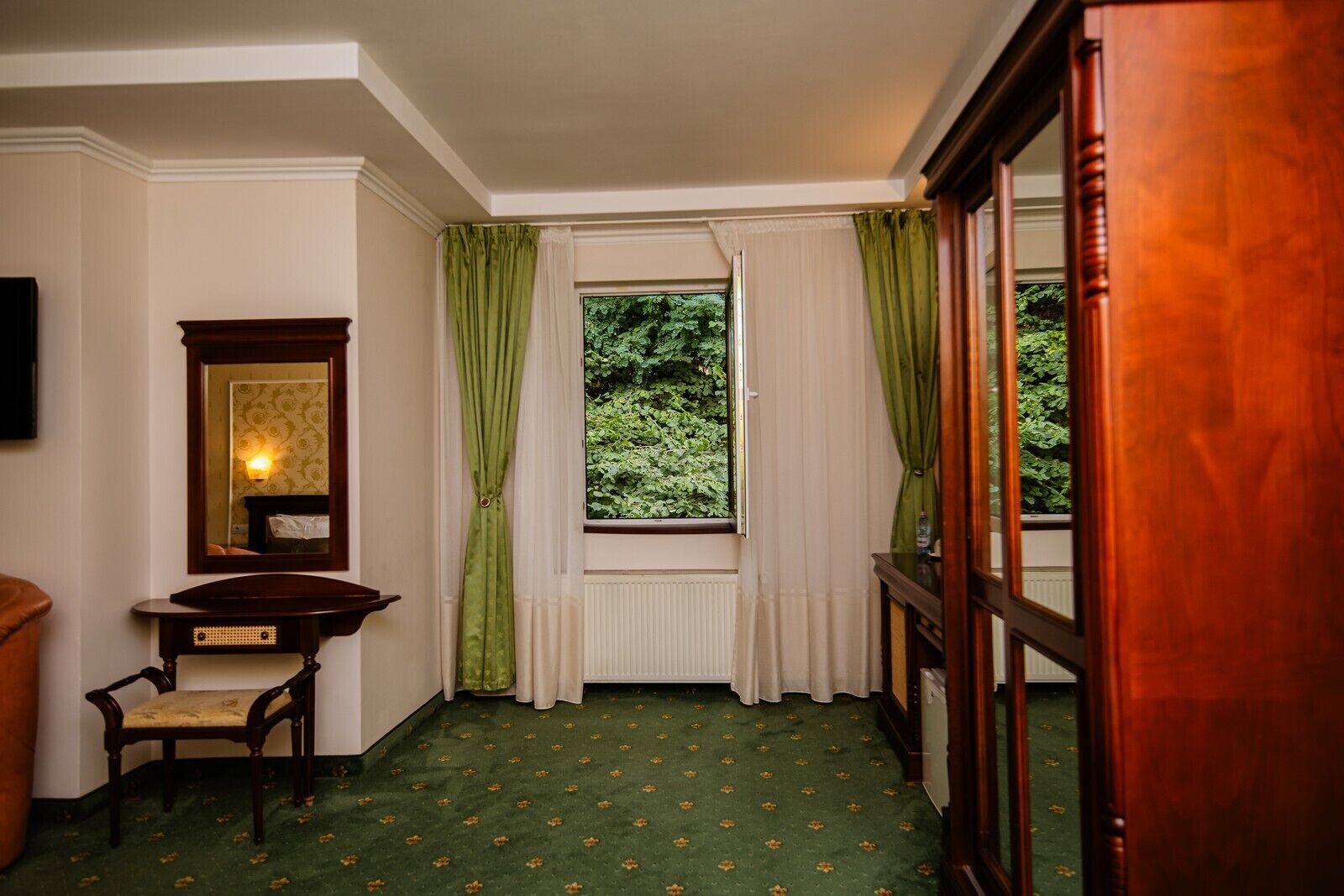 Una habitación con alfombra verde, una silla de madera, un espejo y una ventana con cortinas verdes.