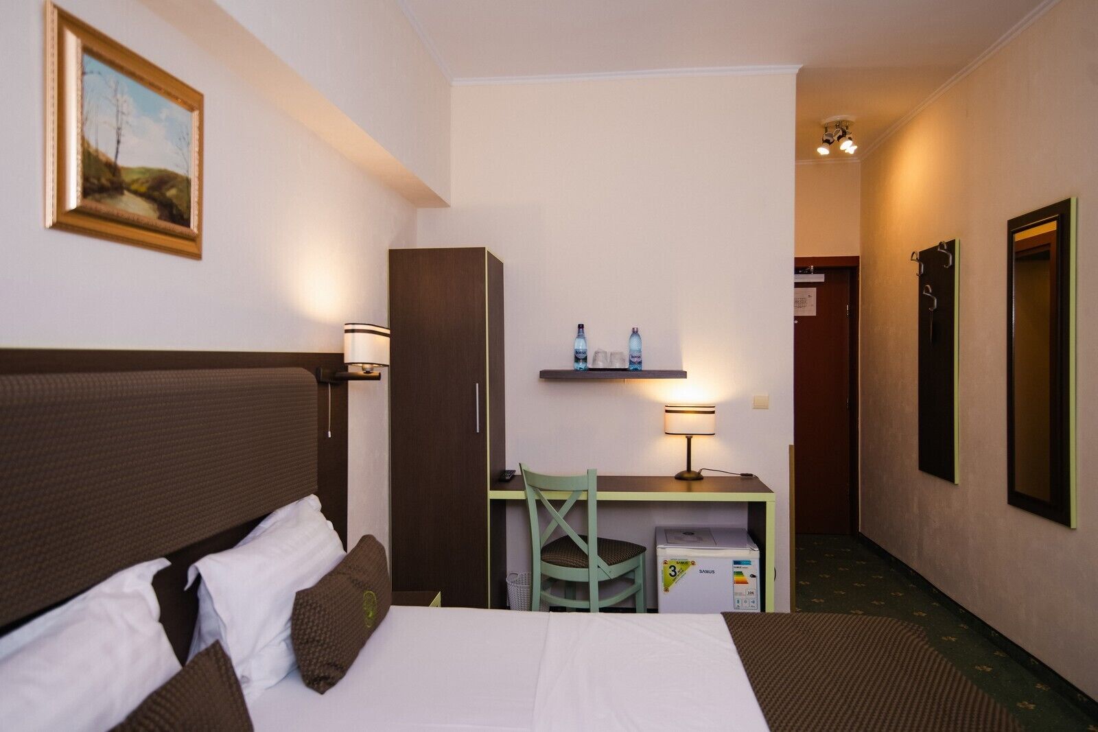 Hotelkamer met een bed, een bureau, een stoel, een lamp, een plank met flessen en een schilderij aan de muur.