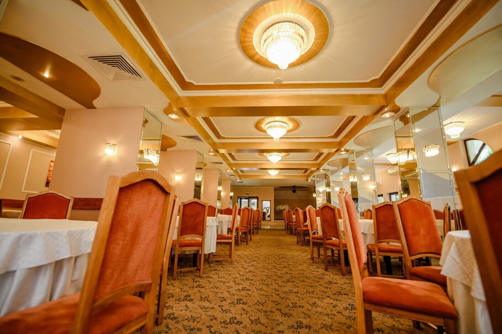 Een restaurantgedeelte in een hotel met rijen tafels en stoelen, kroonluchters en een kamerbreed tapijt met dessin.