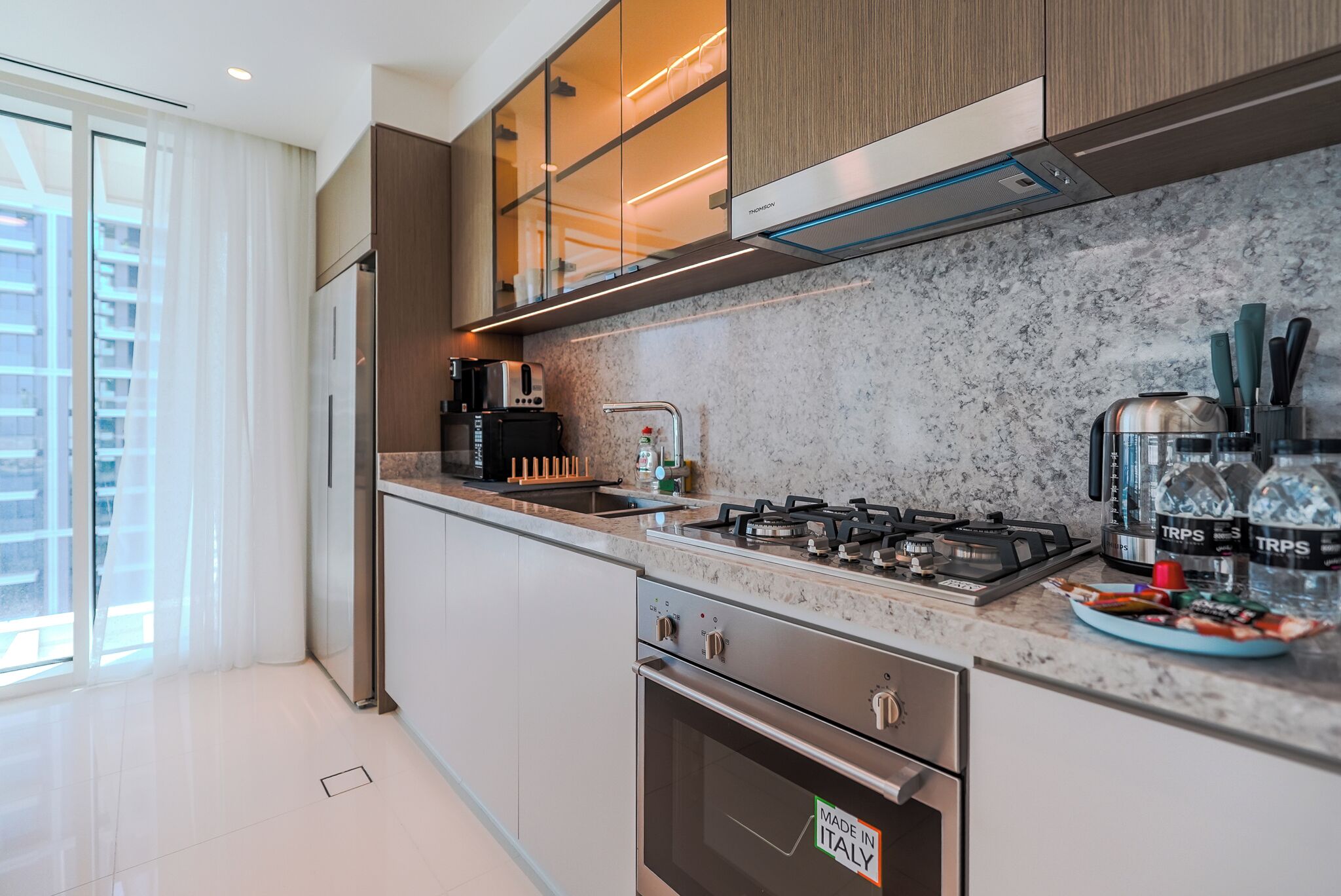 A modern kitchen with stainless steel appliances, a marble countertop, and a view of the city through large windows.