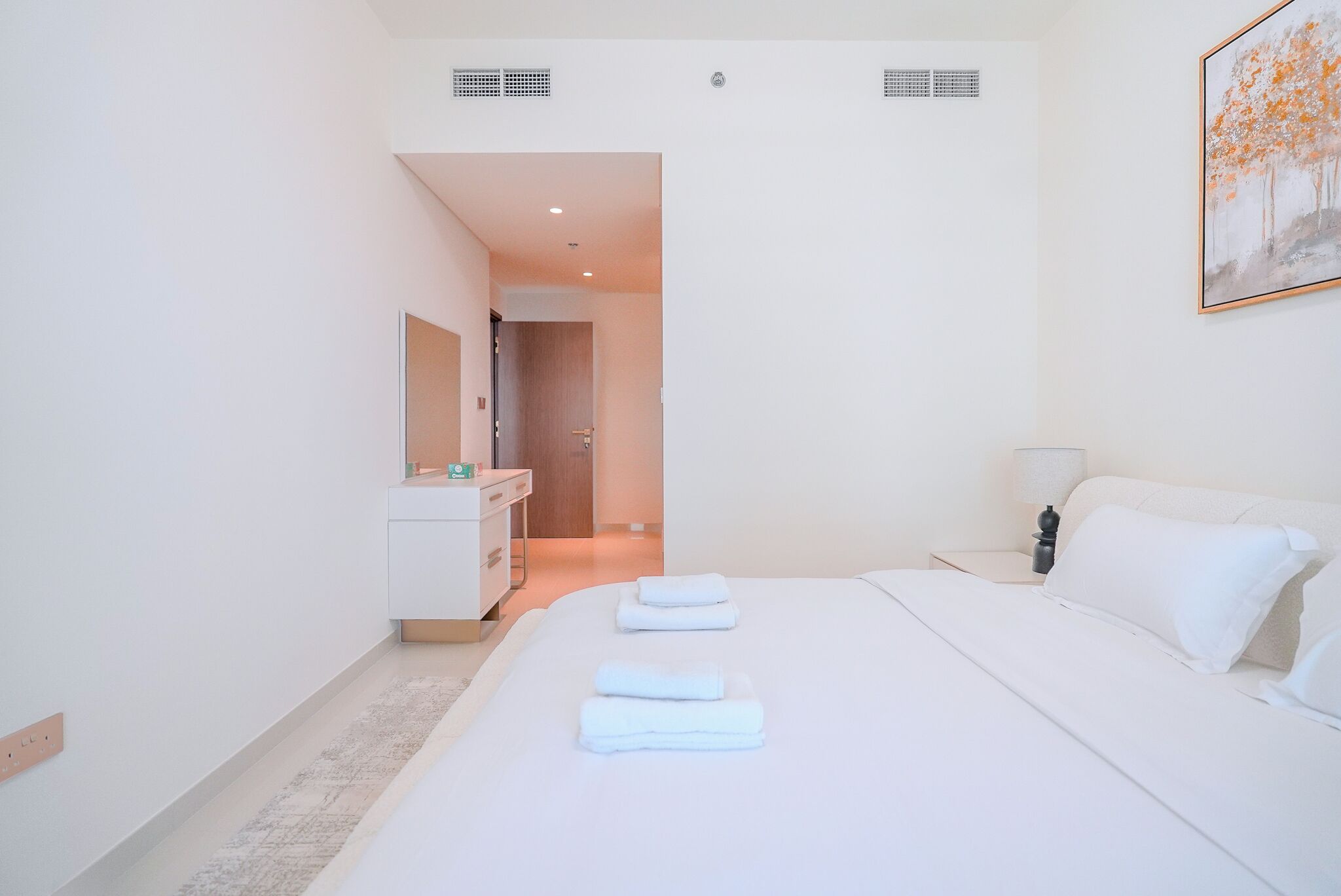 A modern hotel room with a neatly made bed, a bedside table with a lamp, and a bathroom visible through an open door.