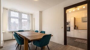A dining area with a wooden table and teal chairs, a window with curtains, and a kitchen visible through an open door.
