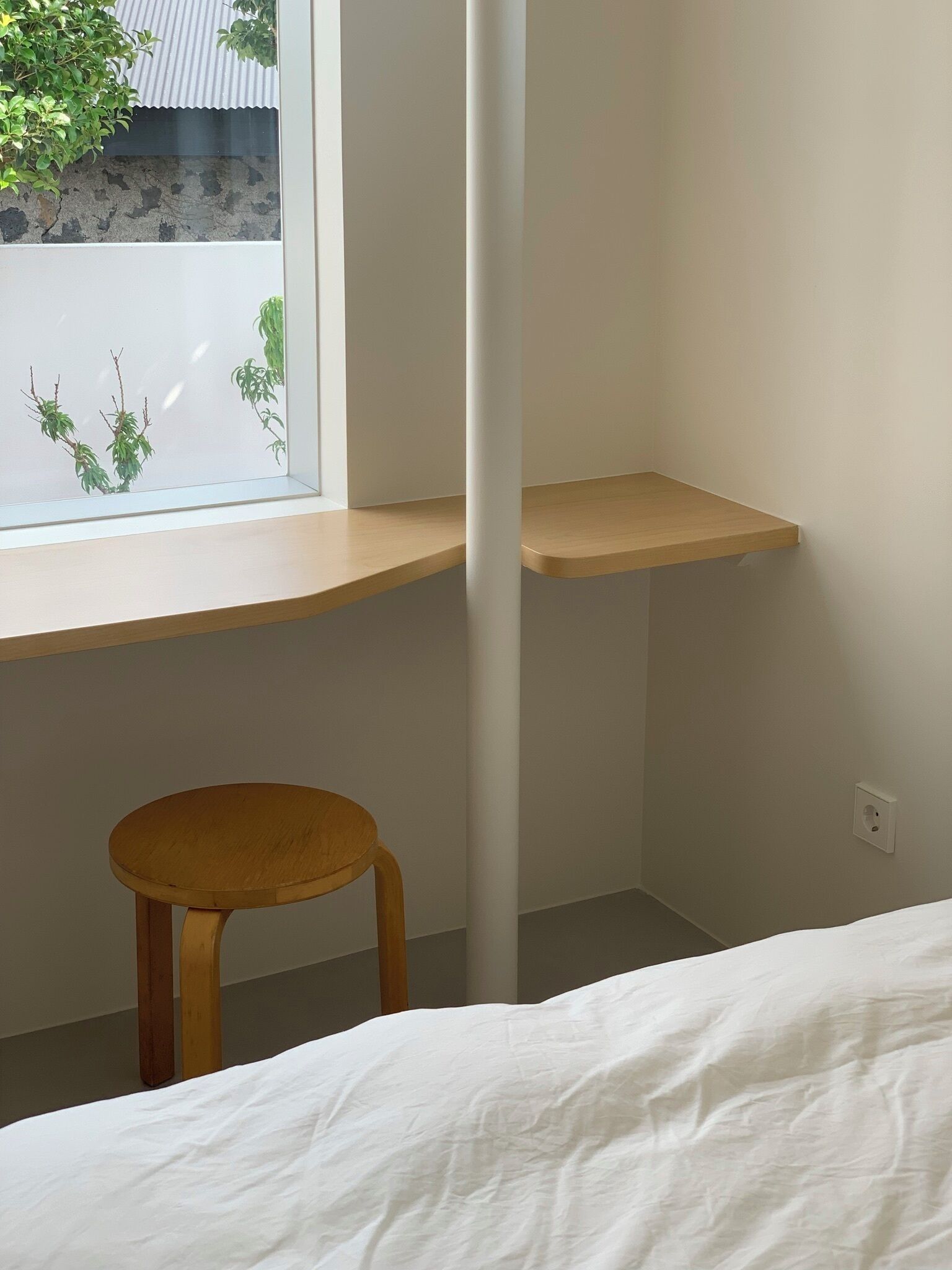 A bedroom with a bed, a wooden stool, a window with a view of a tree and building, and a white wall.