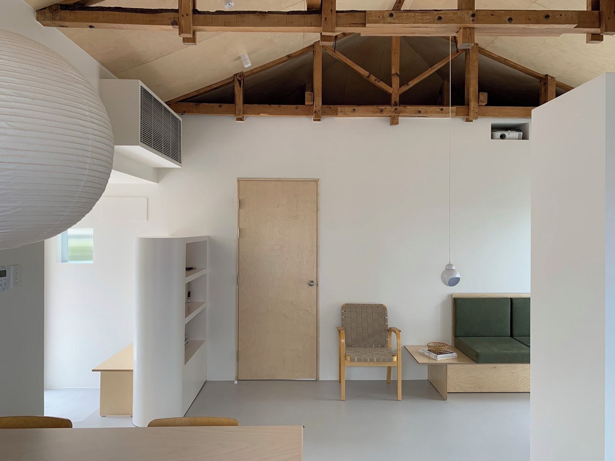 Un interior moderno con techo de madera, una lámpara colgante blanca grande, una puerta de madera, un sofá verde, una silla de madera y una mesa pequeña con una cesta.