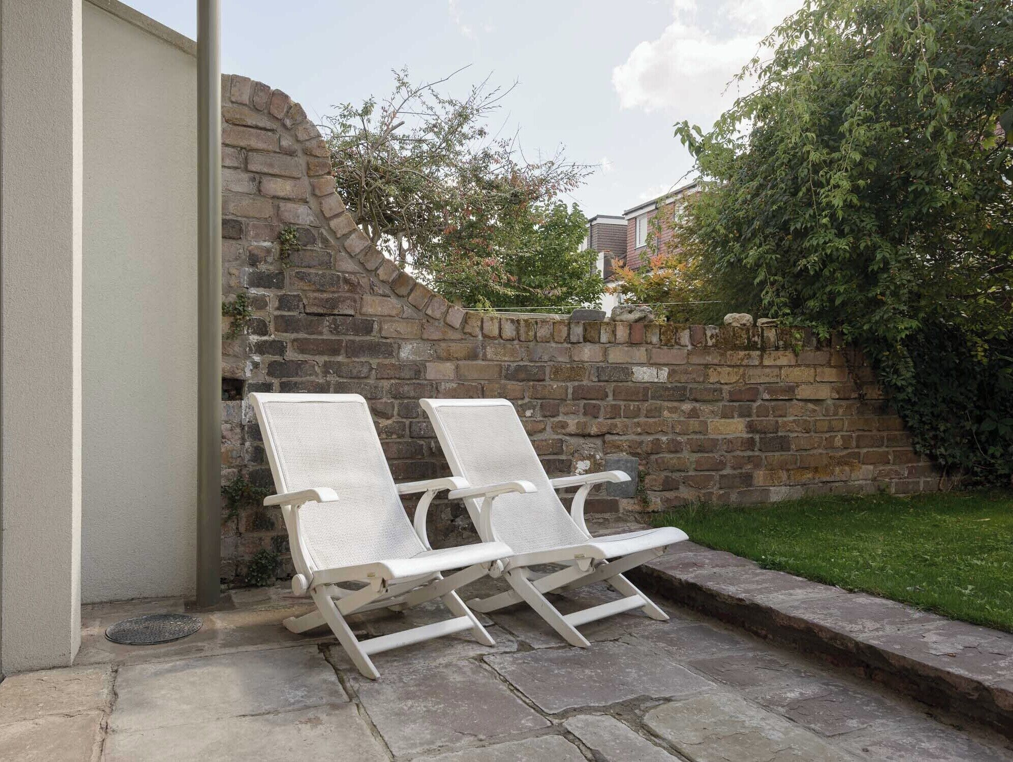 Two white lounge chairs positioned against a stone wall in a garden area.
