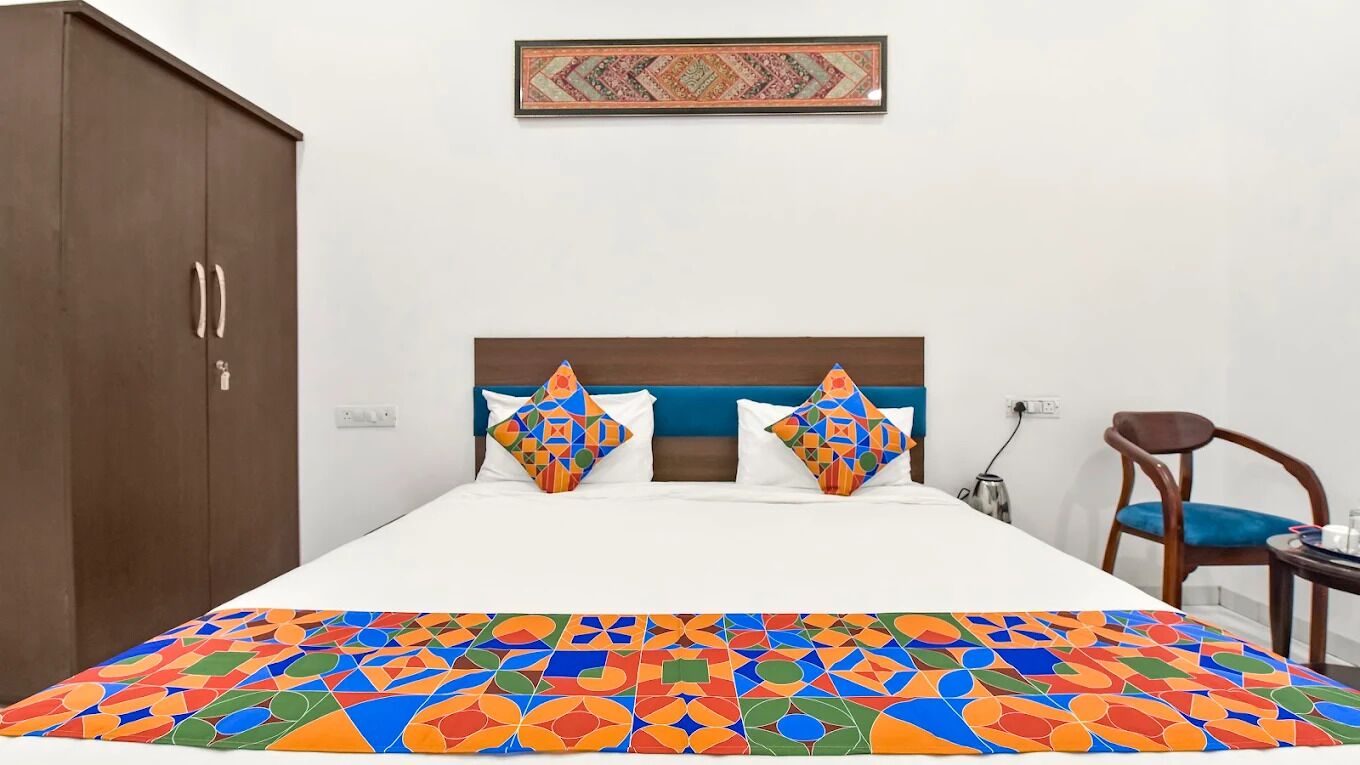 A hotel room with a bed, a wooden wardrobe, a blue chair, and a small table with a kettle.
