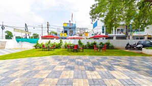Un espace extérieur aménagé avec des chaises rouges et des parasols, un sol pavé, et un bâtiment à l’arrière-plan dont la façade est bleue et jaune.