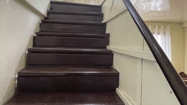 A staircase with dark steps and a black handrail, leading upwards in a narrow corridor with white walls and a window at the top. - Charming 1-bedroom apartment house in Pinamalayan,Oriental Mindoro (Mimaropa)