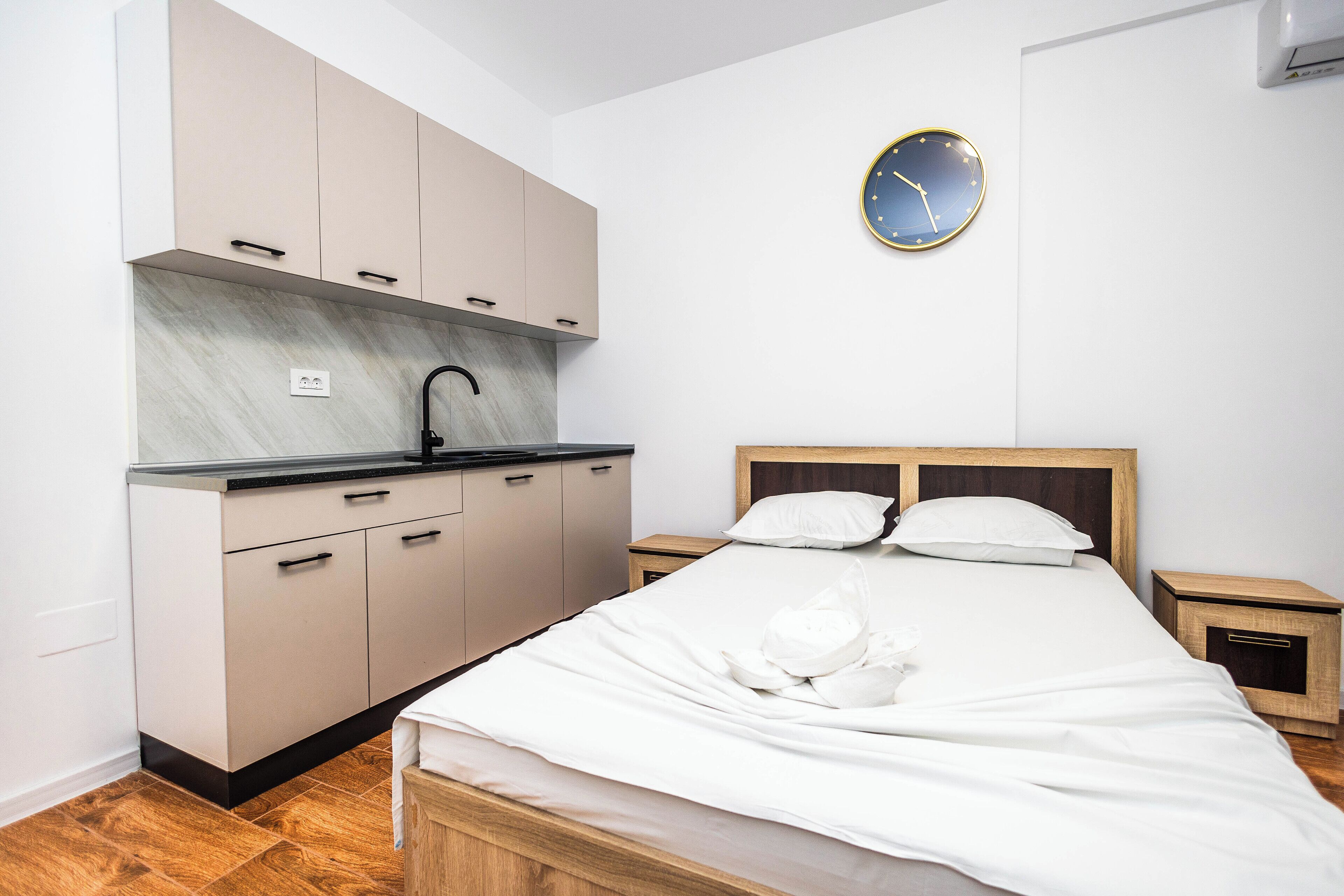 Une chambre à coucher de taille réduite, comprenant un lit, des tables de chevet, une petite lampe de chevet, une horloge murale et un coin cuisine intégré avec un évier et des placards.