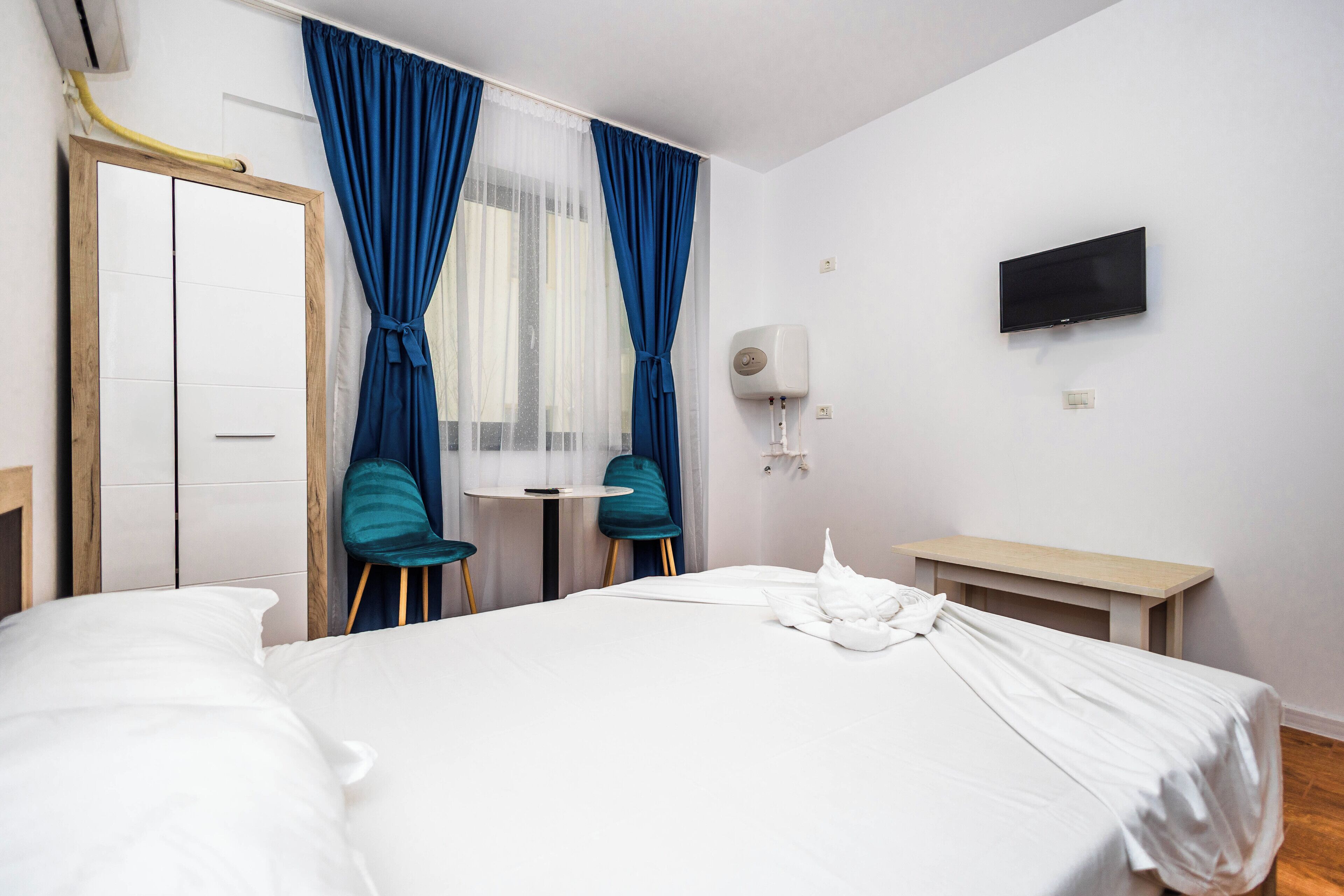 Une chambre d’hôtel avec un lit, deux chaises, une petite table, un téléviseur et une fenêtre avec des rideaux.