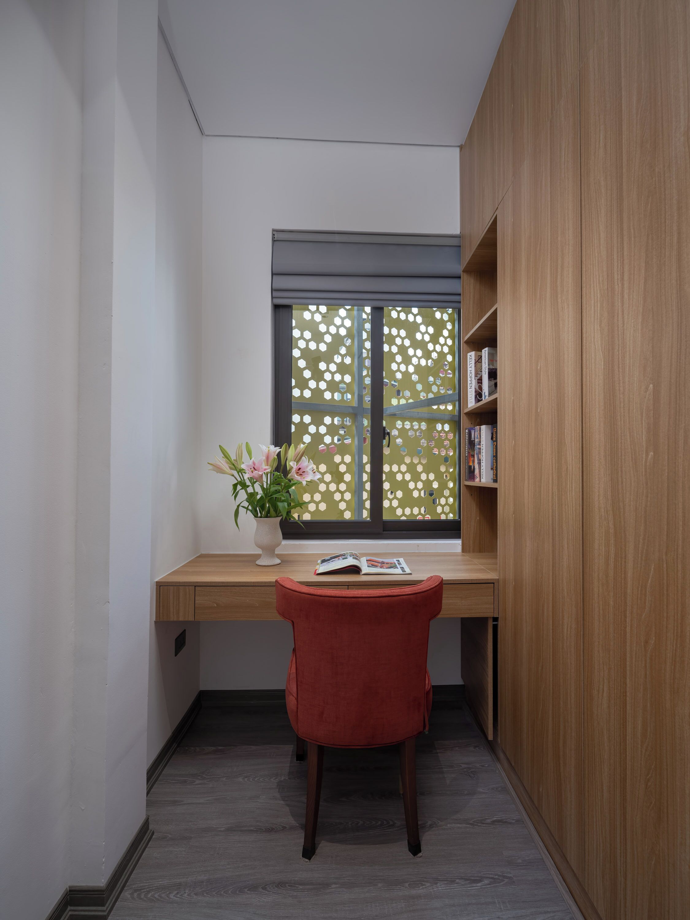 Une pièce d’angle avec un bureau en bois, une chaise rouge, une fenêtre ornée d’un paravent décoratif et une bibliothèque.