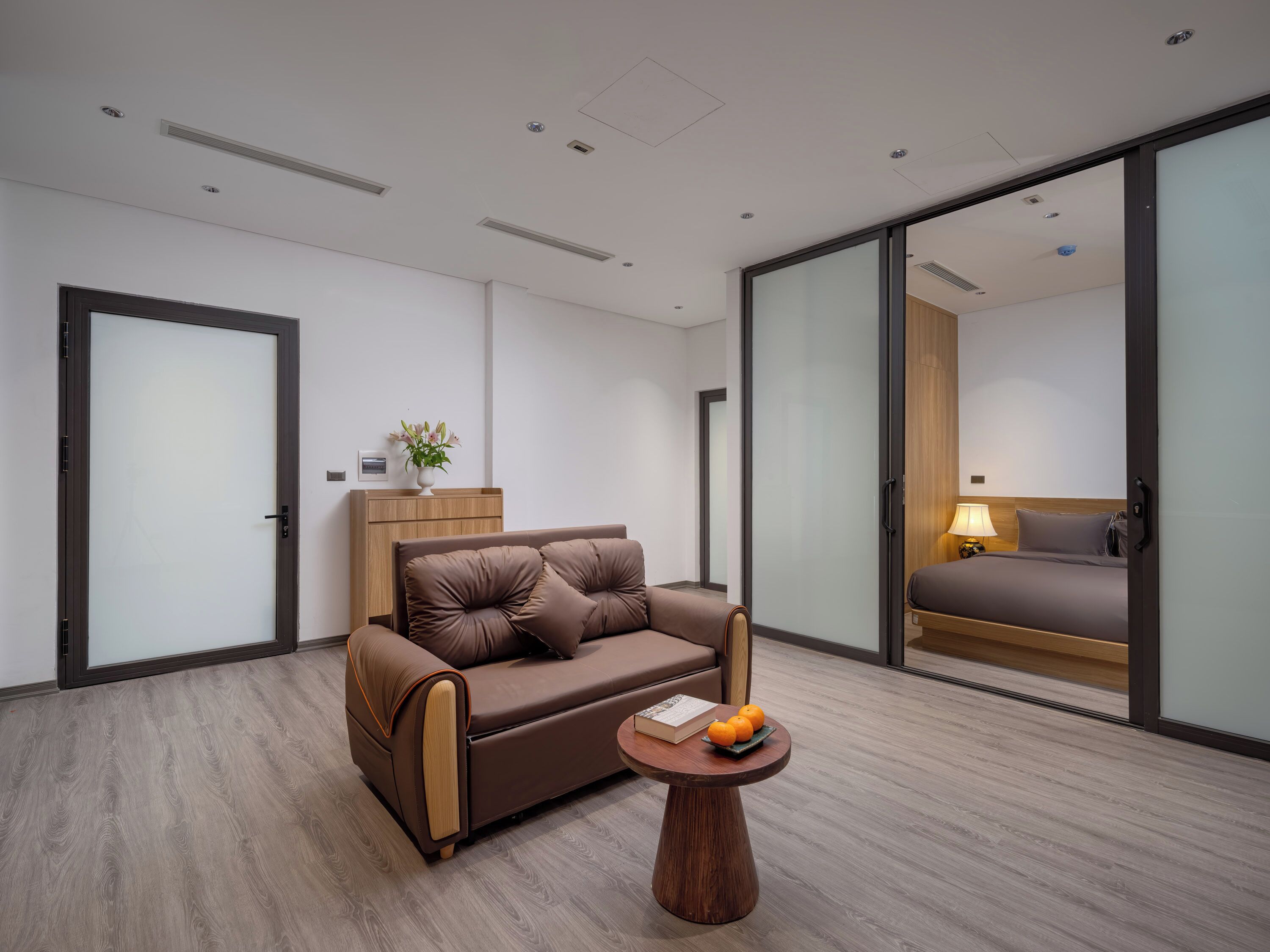 A modern hotel room with a brown leather sofa, a wooden side table with a book and oranges, and a bed with a wooden headboard.