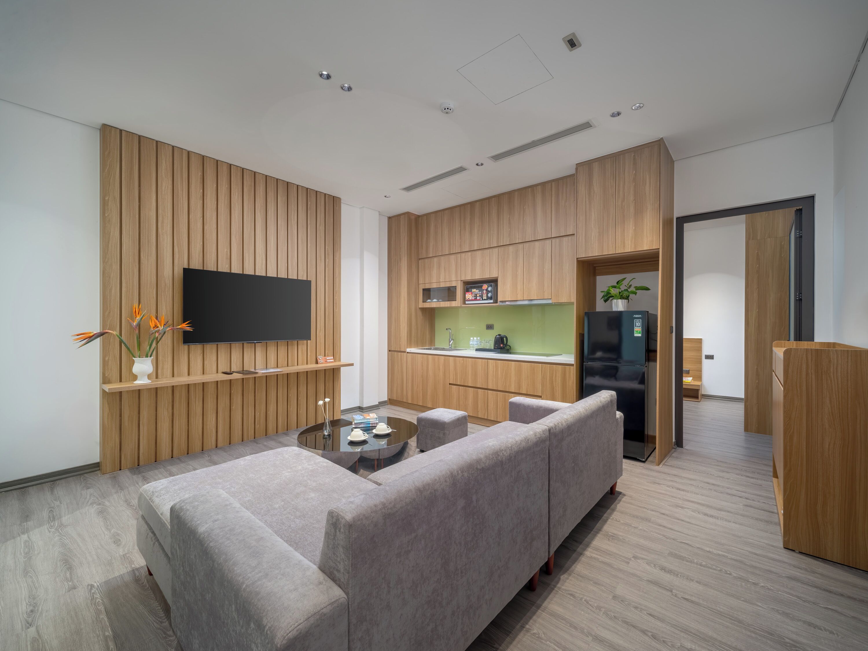 A modern living room with a sofa, coffee table, flat-screen TV, wooden panel walls, and a kitchen area with a refrigerator and sink.