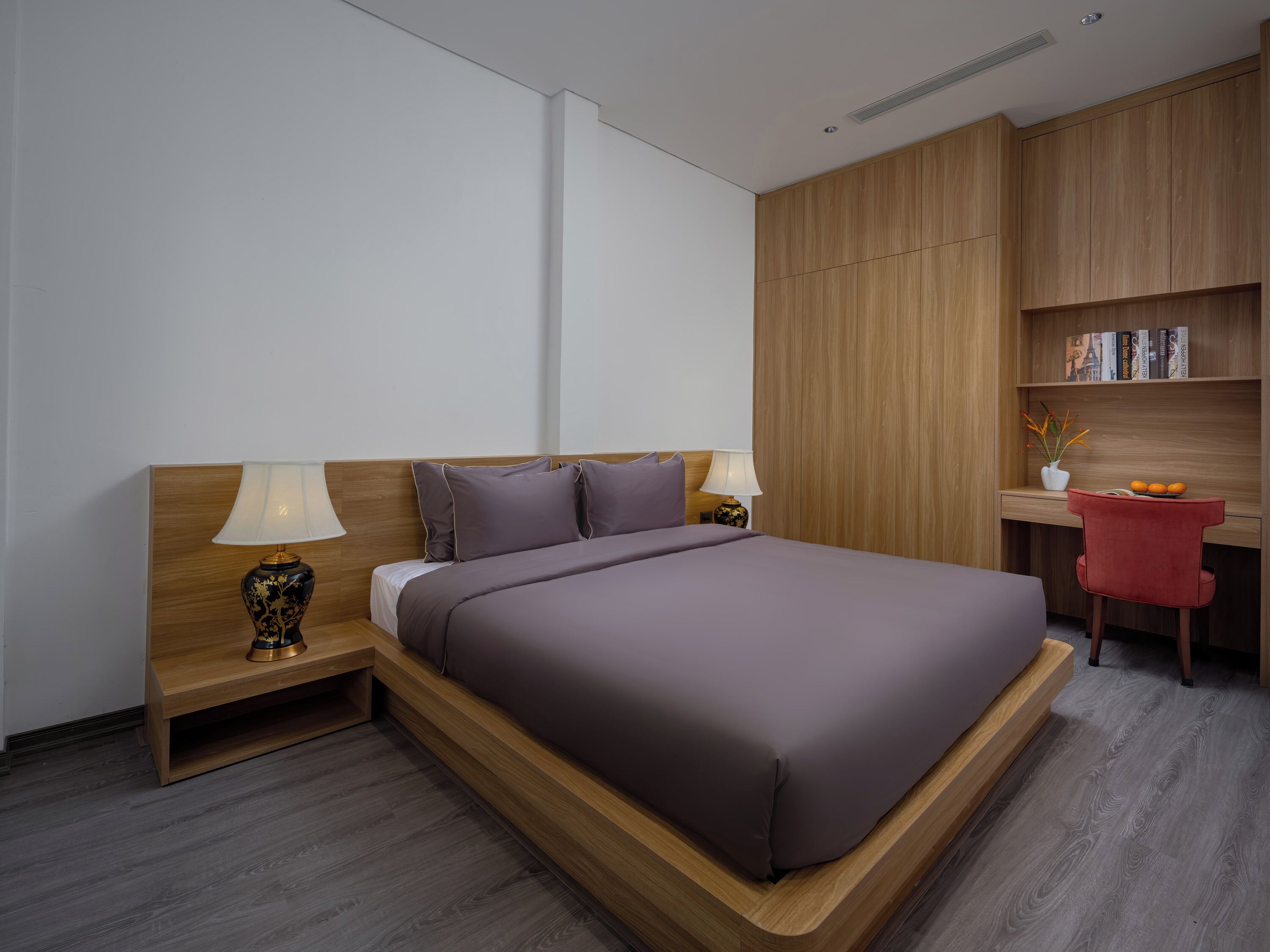A modern hotel room with a large bed, bedside tables with lamps, a desk with a chair, and a bookshelf.