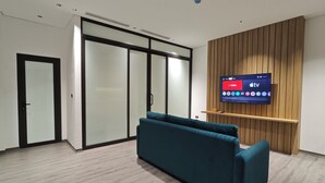 A modern office space with a teal sofa, a large flat-screen TV mounted on a wooden panel wall, and a sliding glass door leading to another room. - Minerva Aparthotel Hanoi (Hanoi)