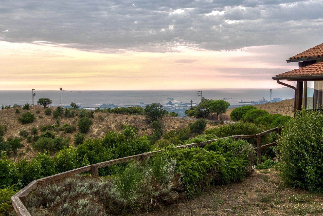 A coastal landscape with a wooden fence, greenery, and a distant view of the sea and ships.