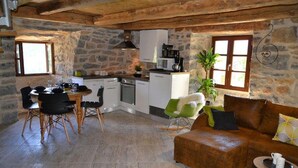 Une cuisine rustique aux murs en pierre, un coin repas avec une table et des chaises, et un espace salon avec un canapé et une petite table d’appoint.