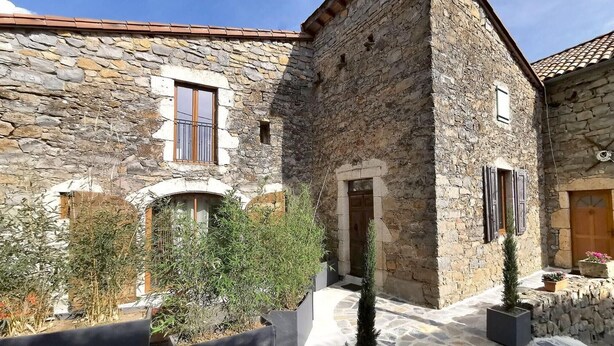 Un bâtiment en pierre doté de fenêtres en arc de cercle et d’une porte, entouré de plantes en pot et d’une cour dallée de pierres.