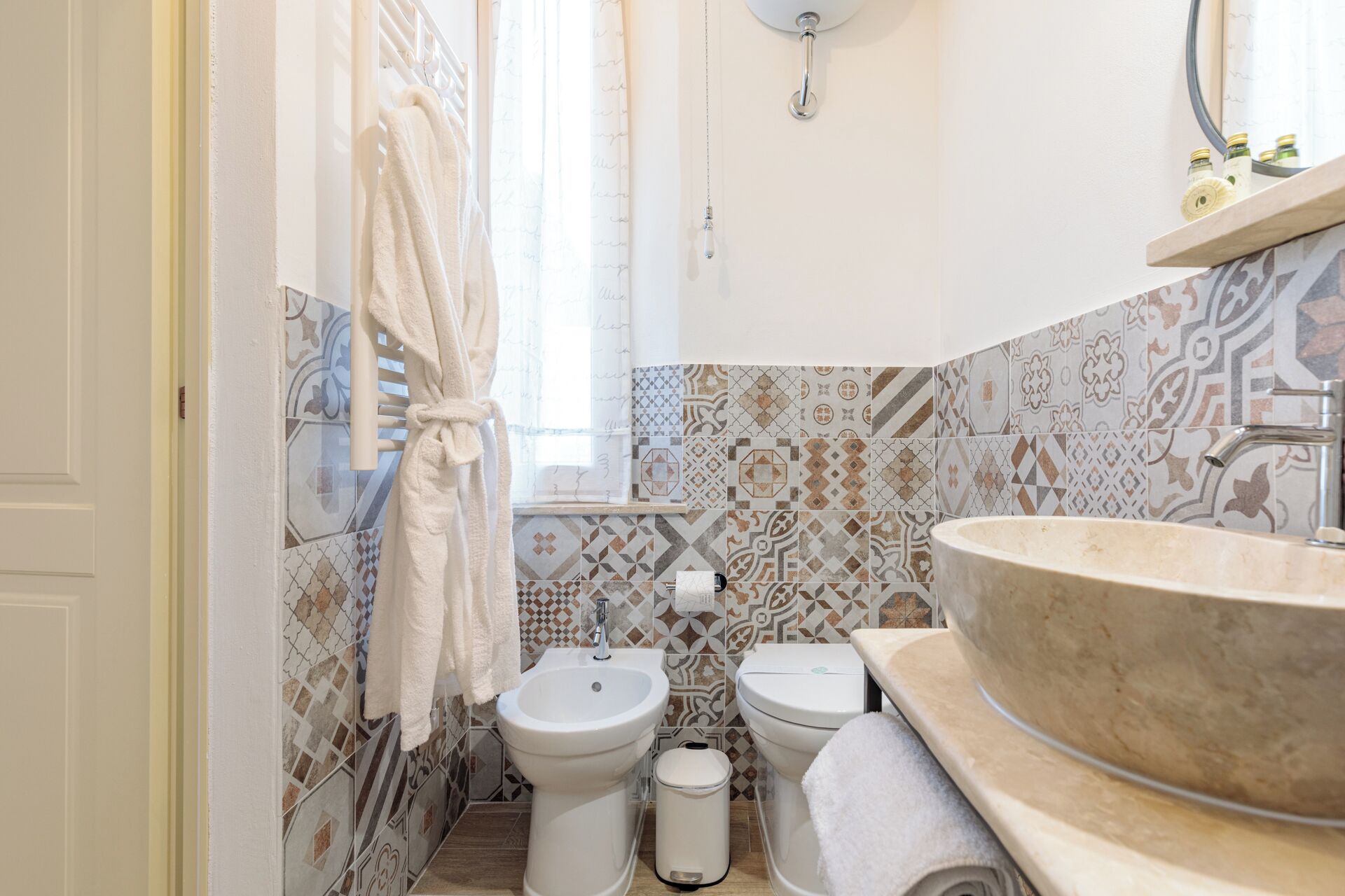 A bathroom with a large sink, two toilets, a bidet, patterned tiles, a towel rack, and a hanging light fixture.