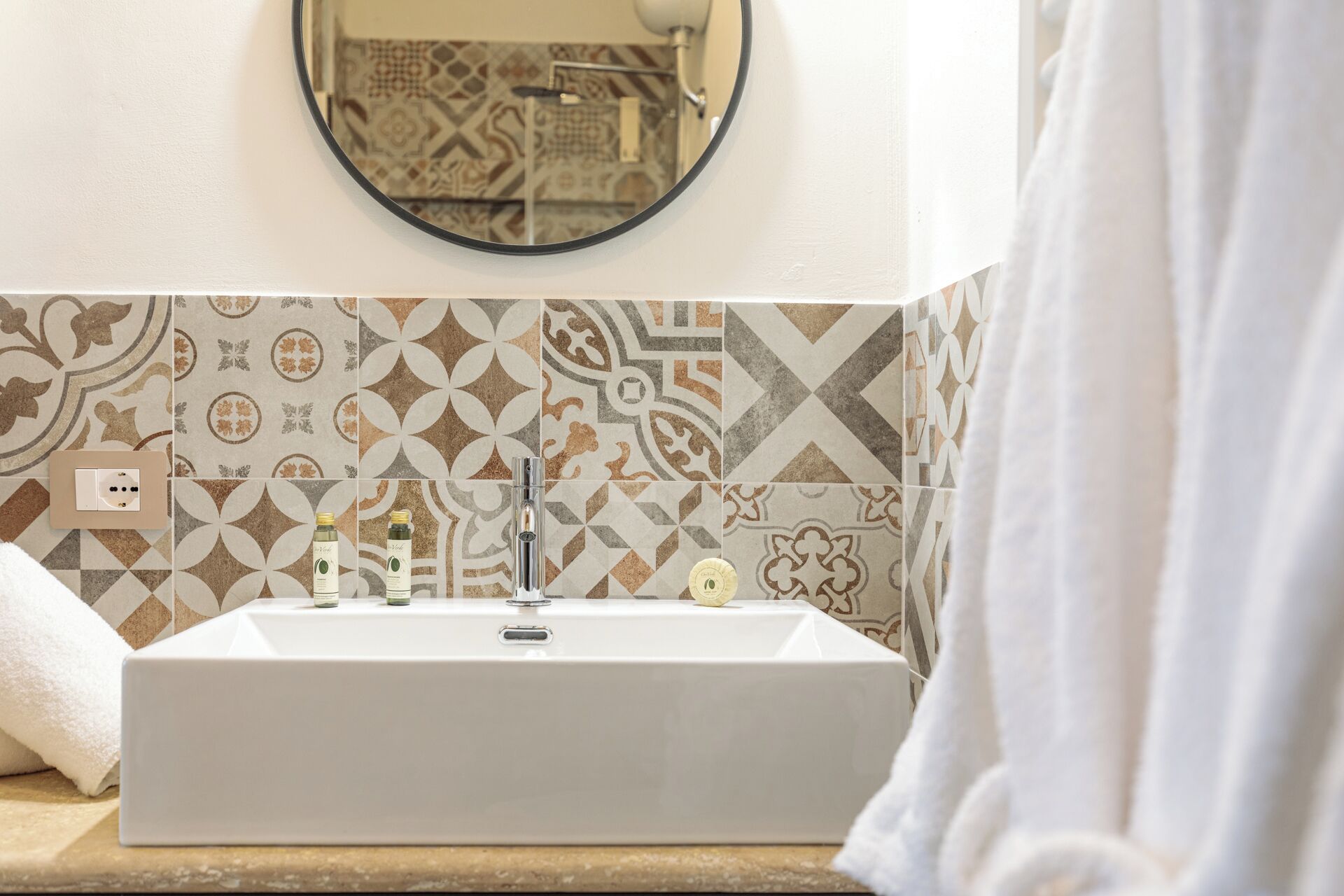 A bathroom with a rectangular sink, a mirror above it, patterned tiles on the wall, and a towel on the floor.