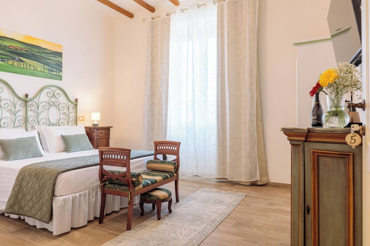 Un dormitorio con cama, dos sillones, una mesita de noche, cómoda, un cuadro y una ventana con cortinas.