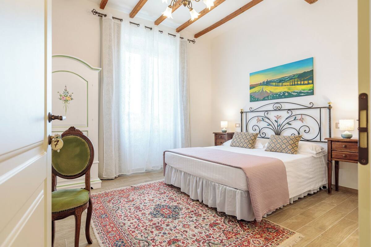 Un dormitorio con cama, mesita de noche, una silla, un cuadro y una puerta con número.