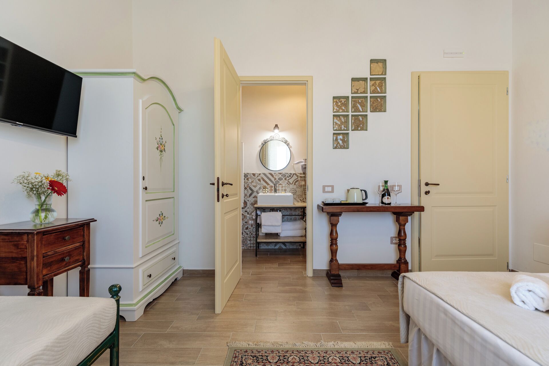 Habitación de hotel con cama, mesita de noche, cómoda, televisión, baño con lavamanos y espejo, y una pequeña mesa con una botella y vasos.