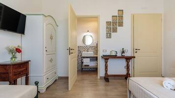 Habitación de hotel con cama, mesita de noche, cómoda, televisión, baño con lavamanos y espejo, y una pequeña mesa con una botella y vasos.