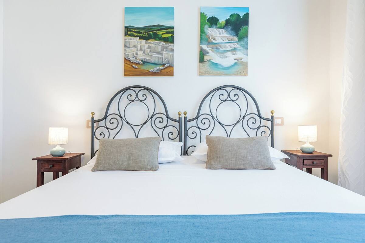 Una cama con cabecera metálica, dos almohadas, mesitas de noche con lámparas y dos cuadros en la pared.