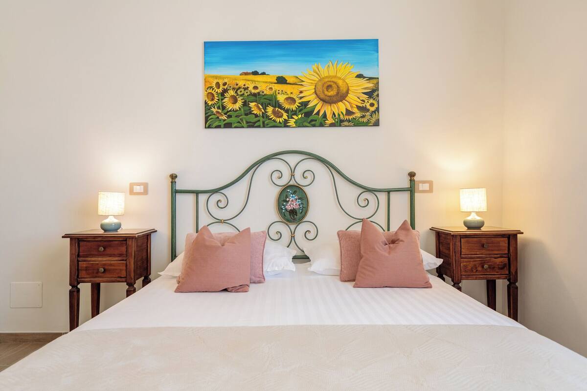 Una cama con cabecera, dos mesitas de noche y un cuadro de girasoles sobre la cama.