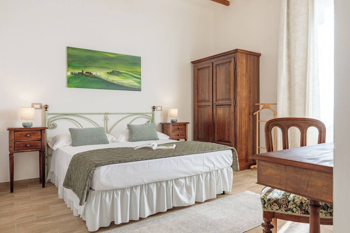 Un dormitorio con cama, mesitas de noche, un armario de madera, una silla y un cuadro en la pared.