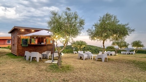 An outdoor seating area with a wooden structure, white plastic chairs, and trees. - Tenuta dell'Argento - Room 7 by Rentbeat (Civitavecchia)