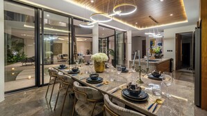 A modern dining area with a long table set for six, surrounded by chairs. The room features a glass partition, a wooden ceiling, and pendant lighting. - Villa Fortuna by RPM (Kerobokan)