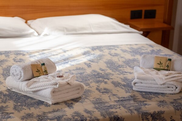 A neatly made bed with a floral-patterned bedspread, two rolled towels with a bottle and a card, and a wooden nightstand with a lamp.