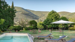 Zona de piscina con sillas de descanso y sombrillas, rodeada de vegetación y colinas.