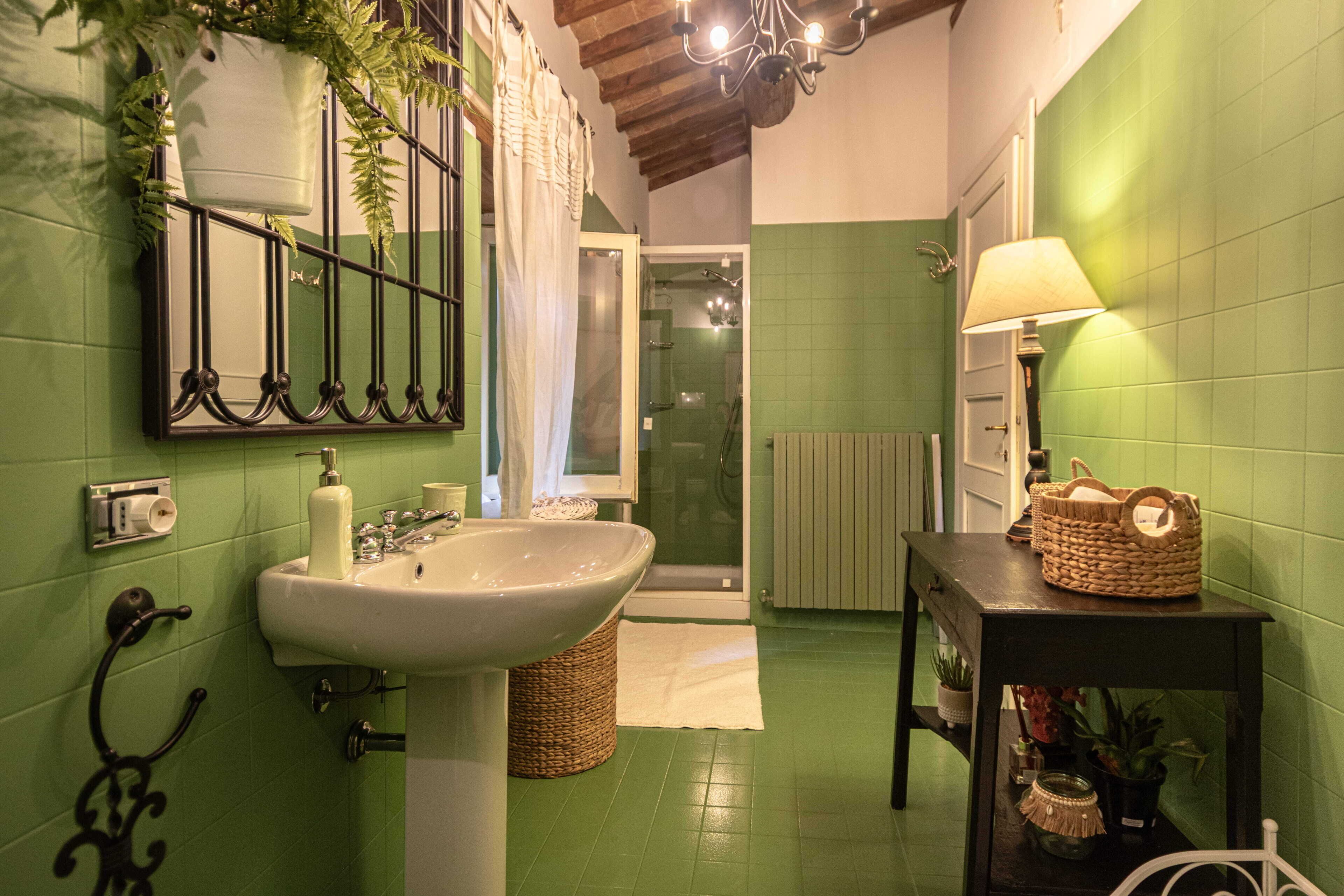 A bathroom with a sink, mirror, shower, and a table with a lamp and basket.