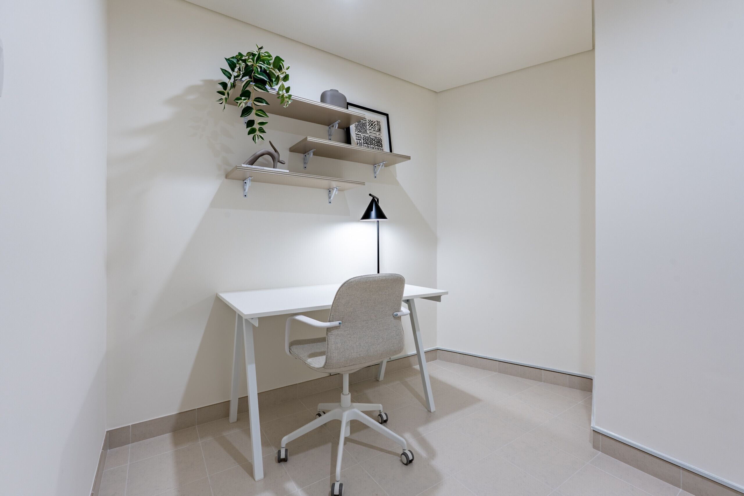 Un bureau blanc avec une chaise, une lampe de bureau noire et une étagère ornée d’une plante et d’objets décoratifs.