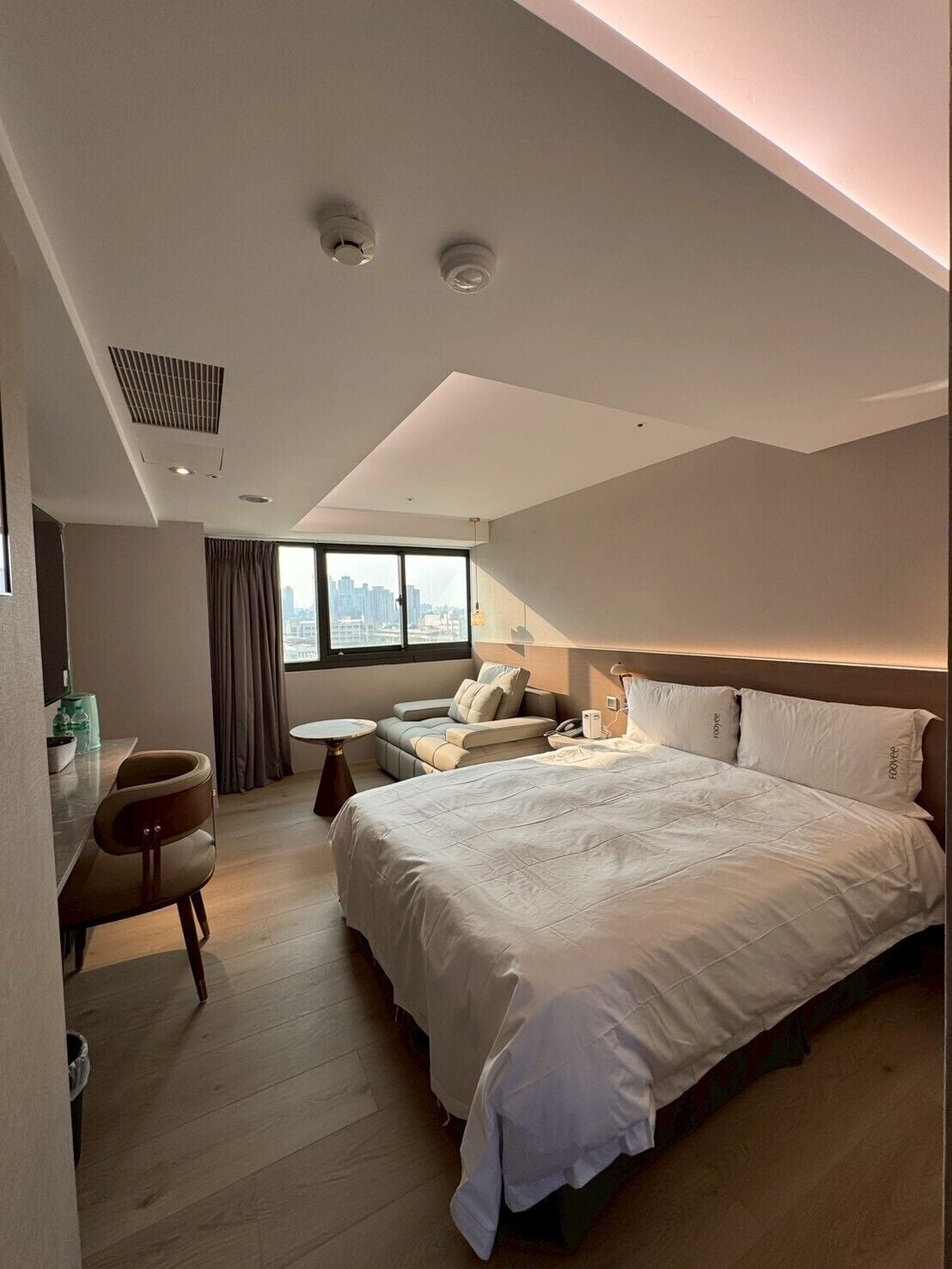 A modern hotel room with a large bed, a seating area with a sofa and armchairs, a small table, and a city view through the window.