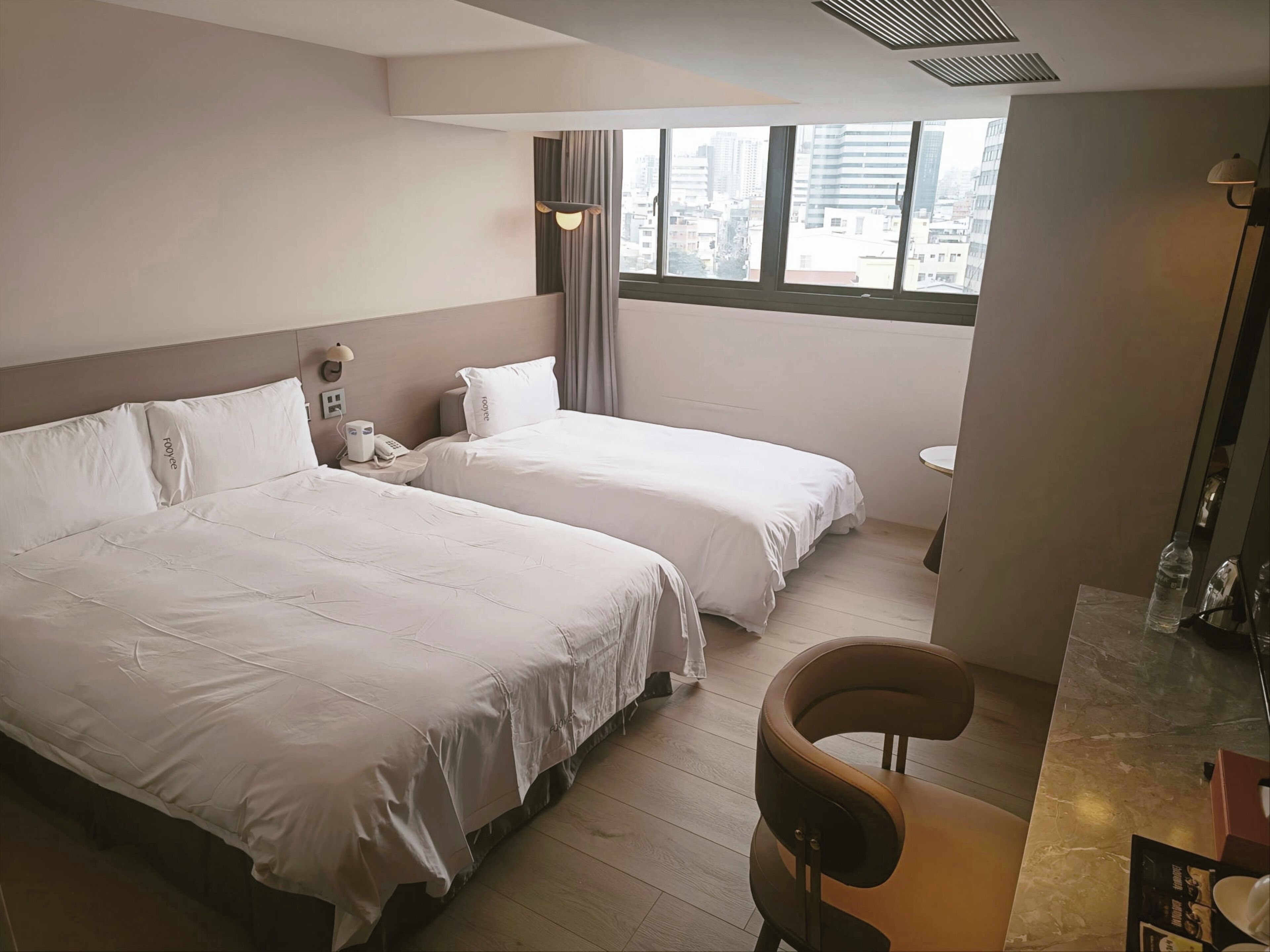 A hotel room with two beds, a small table, a chair, and a view of the city through large windows.