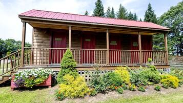 Une cabane en rondins avec un toit rouge, une véranda en bois et un jardin où poussent différentes plantes et fleurs.