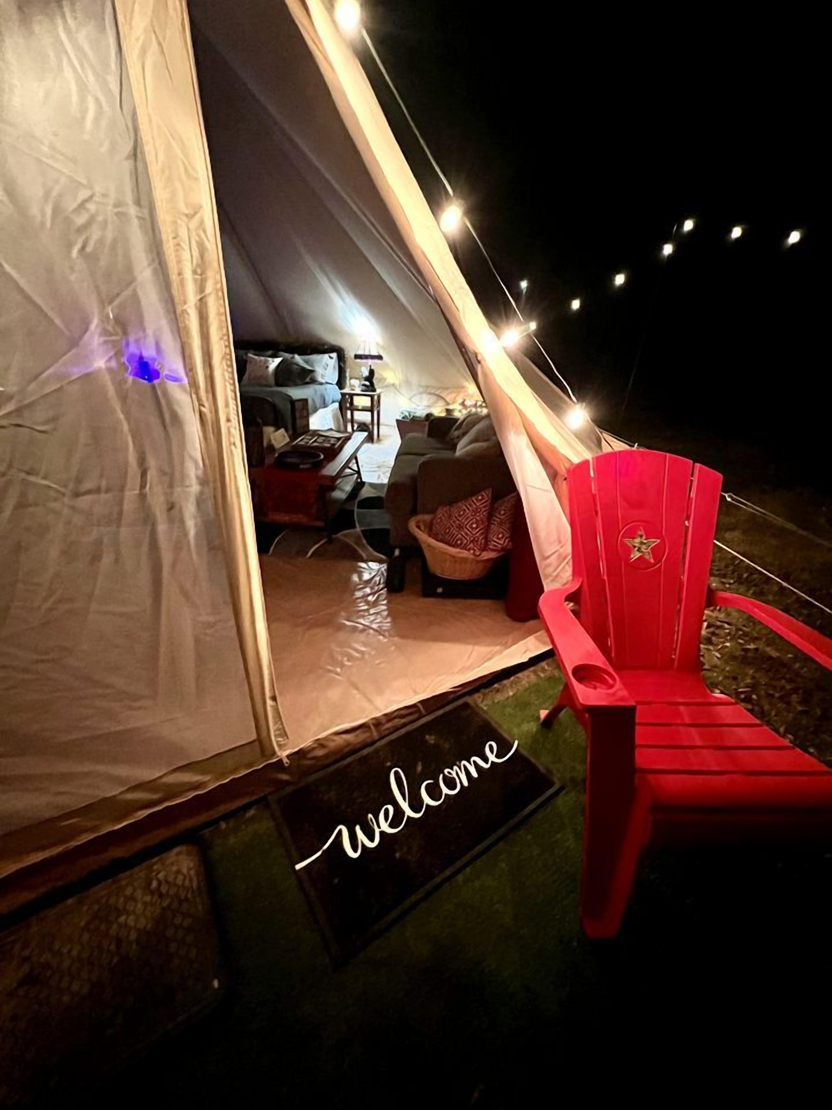A tent interior with a red chair, a welcome mat, and a table with a lamp.