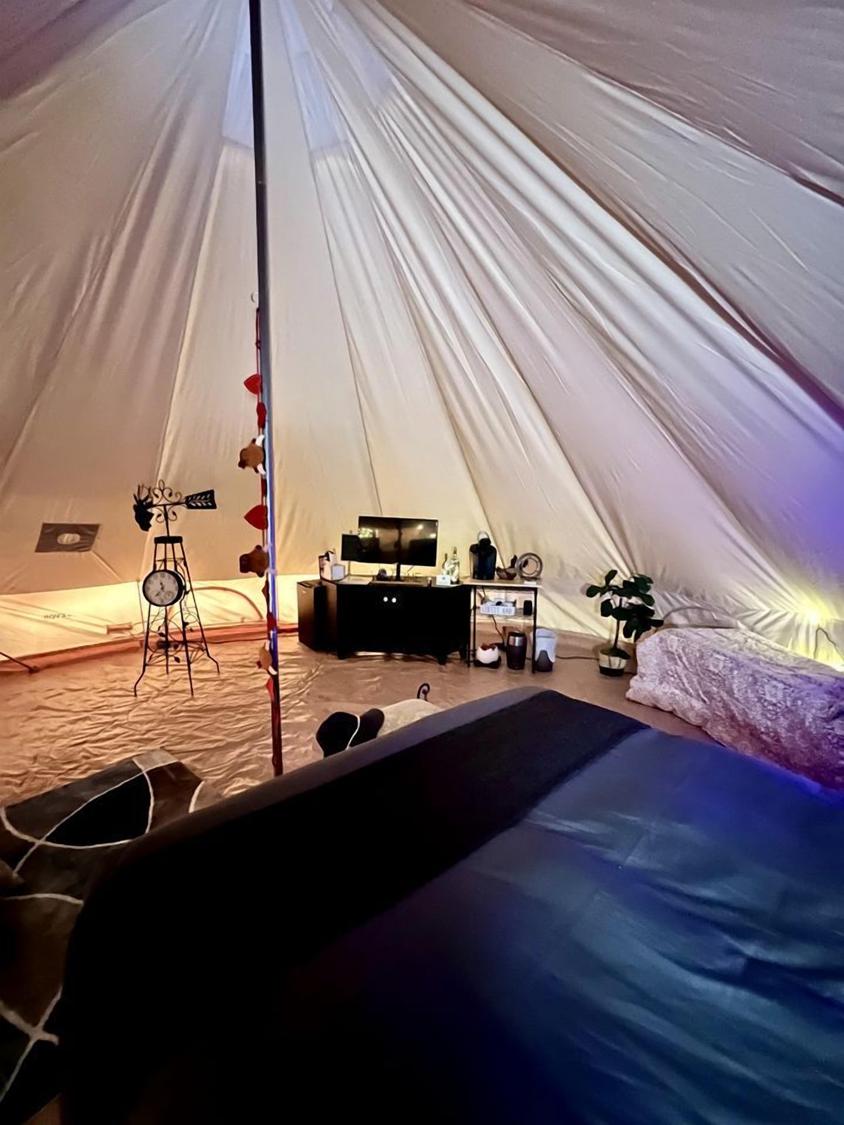 A tent with a bed, a small table, a clock, and a plant.
