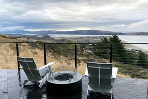A balcony with two white chairs and a circular fire pit, overlooking a scenic landscape with hills and a body of water.