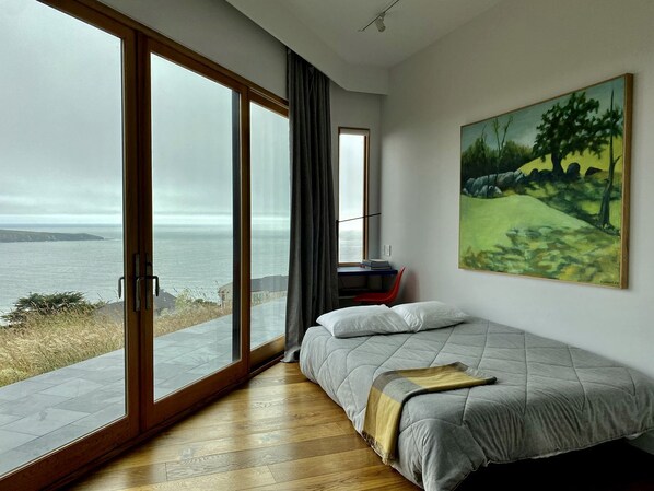 A bedroom with a large sliding glass door leading to a view of the sea, a bed with a blanket, and a painting on the wall.
