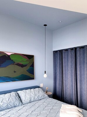 A bedroom with a bed, a hanging light fixture, a painting on the wall, and curtains.